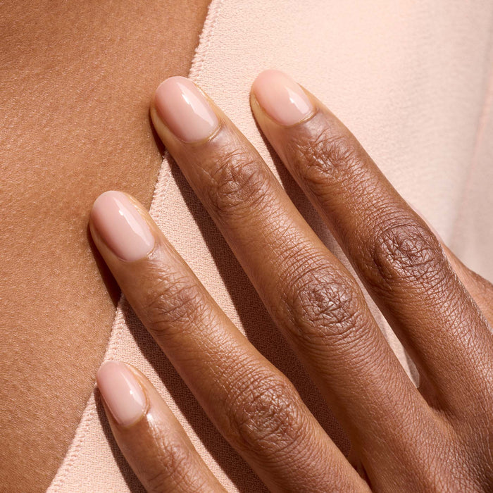 A close-up of a hand with medium-brown skin touching soft light pink fabric. The natural, neatly shaped fingernails are painted with Manucurist’s Pale Rose, a shiny sheer pastel pink gel polish.
