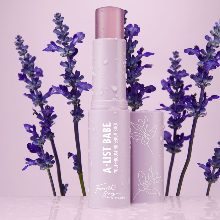 A-List Babe Serum Stick standing uncapped with dewy droplets on a pastel lavender tube, framed by purple lavender sprigs on a glossy pink surface; ColourPop Serum Stick by Fourth Ray Beauty for easy swipe-on face serum.