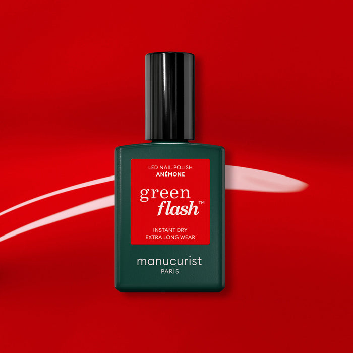 A bottle of Manucurist Anémone gel polish stands out on a bold red background with a white brushstroke. This light red shade from Manucurist Paris pops with vibrant color and shine.