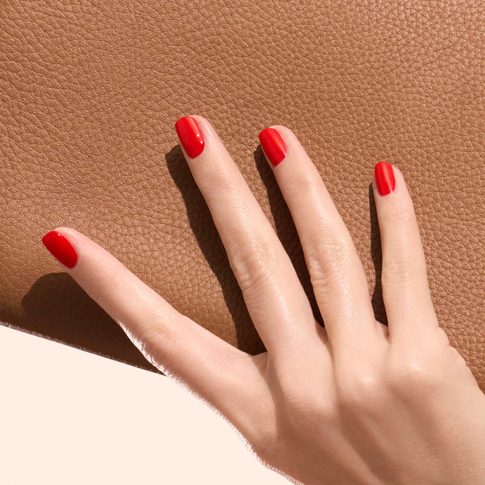 A hand with neatly manicured nails painted in Manucurist’s Anémone, a vibrant red shade, rests on a textured brown surface against a soft, neutral background.