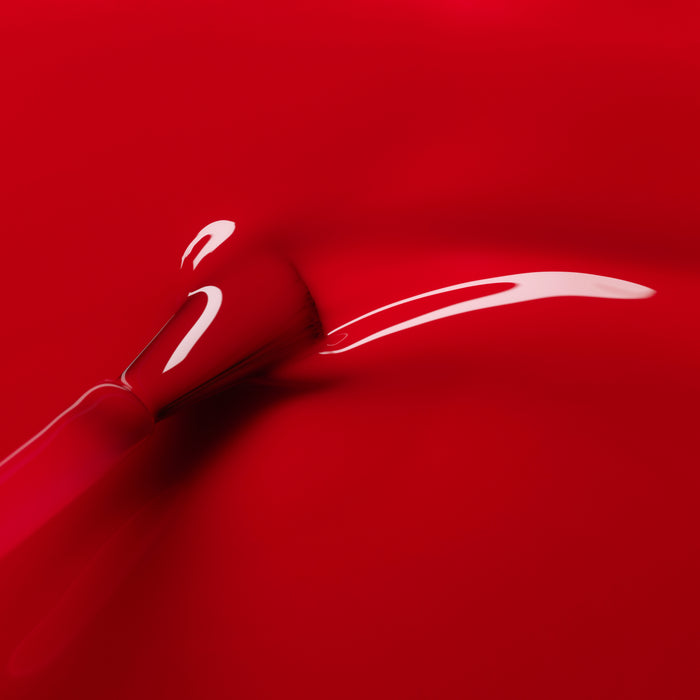 A close-up of a brush applying Manucurist's Anémone nail polish, creating a smooth, shiny red streak on a vibrant background.