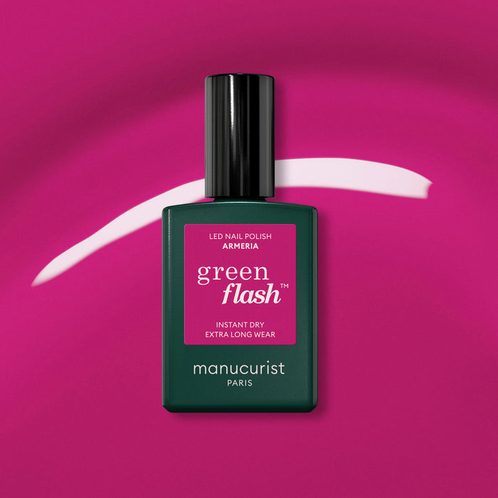 A bottle of Manucurist Armeria rests on a vibrant pink background with a curved mauve streak behind it. The dark green bottle features a black cap and a bright pink label.