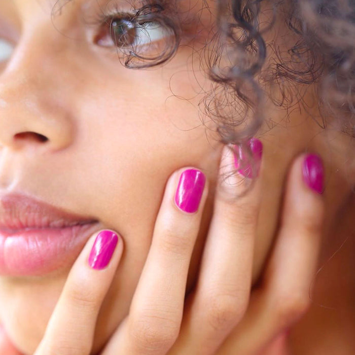 A close-up of a woman with natural skin and curly hair gently touching her cheek, showcasing shiny mauve Armeria gel polish by Manucurist on her fingernails.