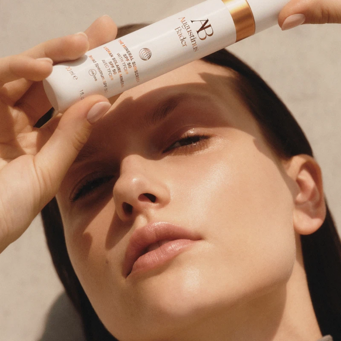 Augustinus Bader The Mineral Sunscreen SPF 50 tube held above a woman's face