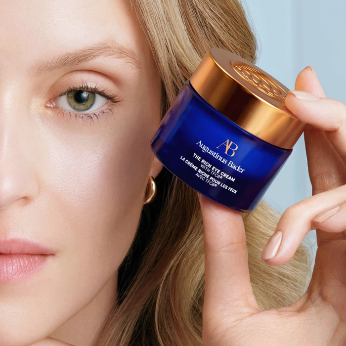 Augustinus Bader The Rich Eye Cream blue jar with gold lid shown beside a woman's eye