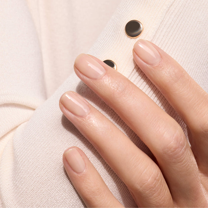 A close-up of a hand with neatly manicured nails painted in Manucurist's Beige, a sheer nude shade, resting on light beige fabric with two gold and black buttons.