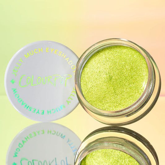 ColourPop Jelly Much Shadow in Billionaire sits open, revealing a metallic lime-green gel shimmer; the white lid with holographic Jelly Much Eyeshadow text rests beside it on a mirrored surface against a soft pastel gradient backdrop.