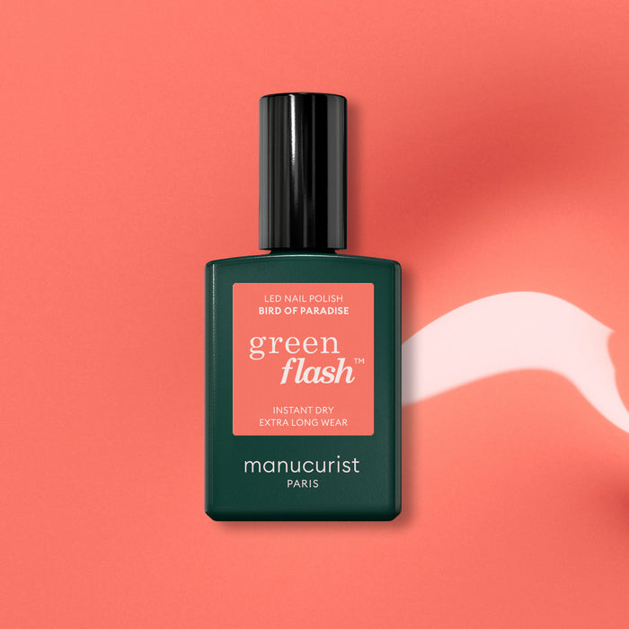 A vibrant coral orange gel polish, Manucurist Bird of Paradise, is displayed in a green bottle on a coral background with a white paint-like streak beside it.