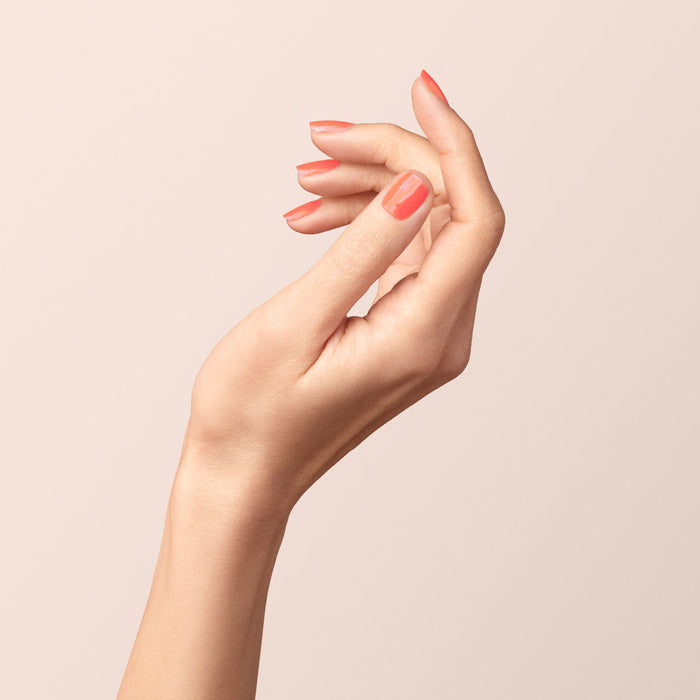 A close-up of a raised hand with neatly manicured nails painted in Manucurist’s Bird of Paradise, a glossy coral orange gel polish, against a light beige background.