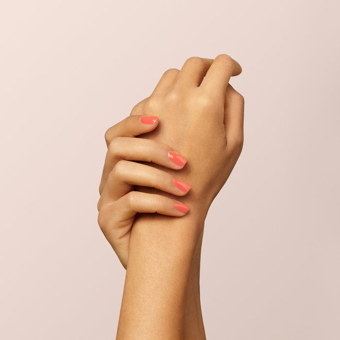 A close-up of two hands crossed gracefully against a light beige background, featuring neatly manicured nails painted with Manucurist’s Bird of Paradise, a glossy coral orange gel polish.