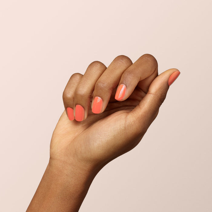 A hand with medium brown skin shows neatly manicured nails painted in Manucurist’s Bird of Paradise, a glossy coral orange gel polish, against a light background.