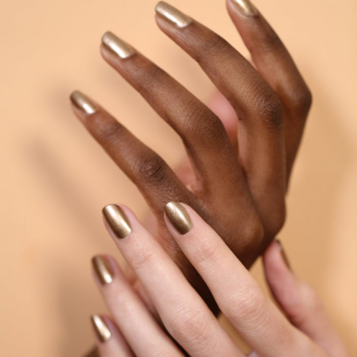 Two hands with varied skin tones, each sporting short, manicured nails painted in Manucurist’s Bronzé metallic gel polish, are beautifully posed against a soft beige background.