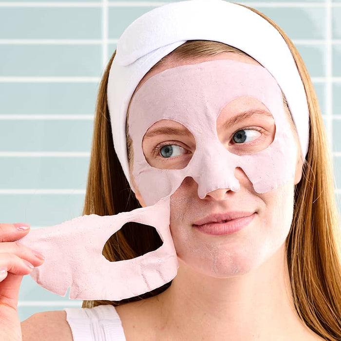 Woman peeling off pink sheet mask from face, soothing skincare treatment against tiled background