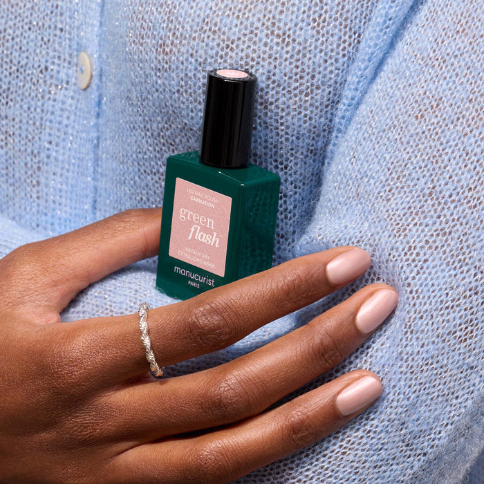 A person in a light blue knit sweater holds a bottle of Manucurist Carnation gel polish. Their nails are manicured in a pearly pink nude shade, accented by a delicate silver ring on their finger.