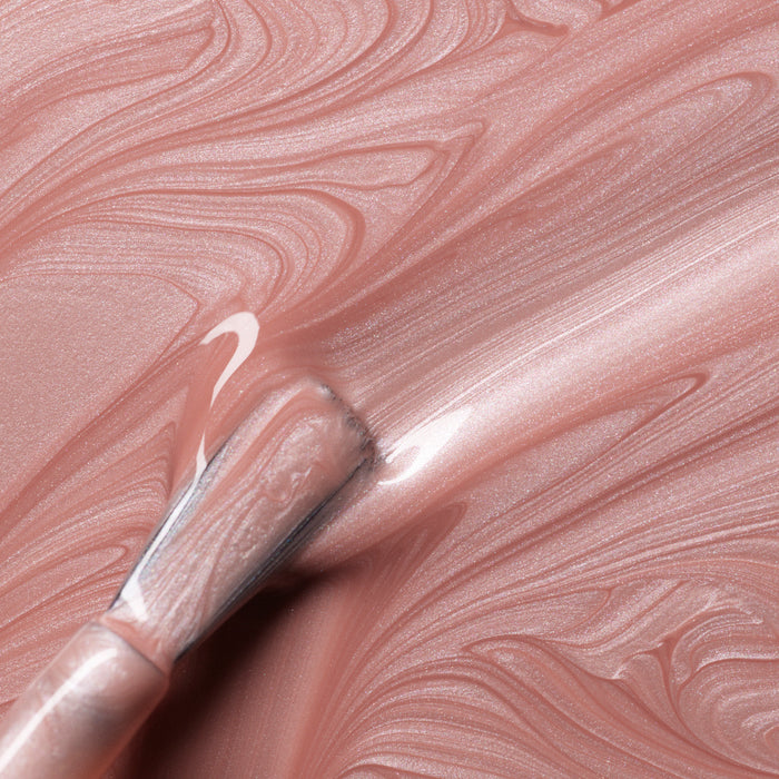A close-up of a brush applying Carnation by Manucurist—a shimmery, rose gold gel polish—over a marbled pearly pink surface, creating smooth, glossy streaks in the Green Flash formula.