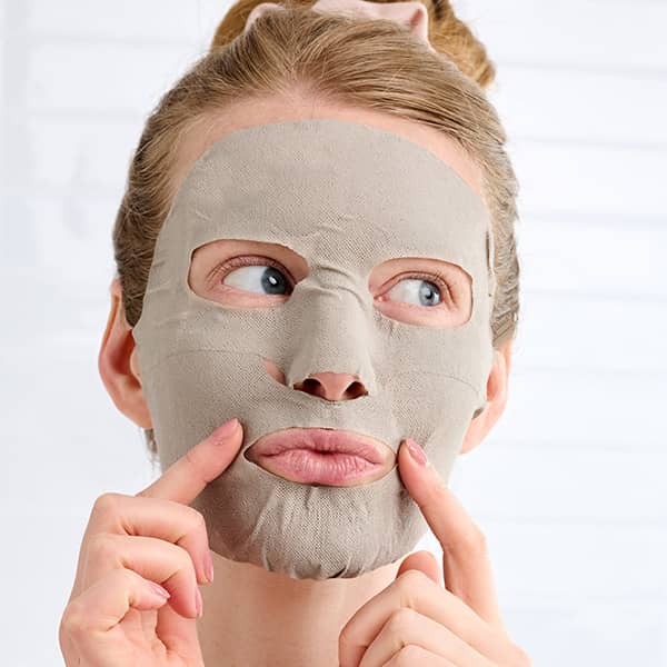 Woman wearing beige sheet face mask, pressing cheeks with fingers while relaxing during skincare treatment