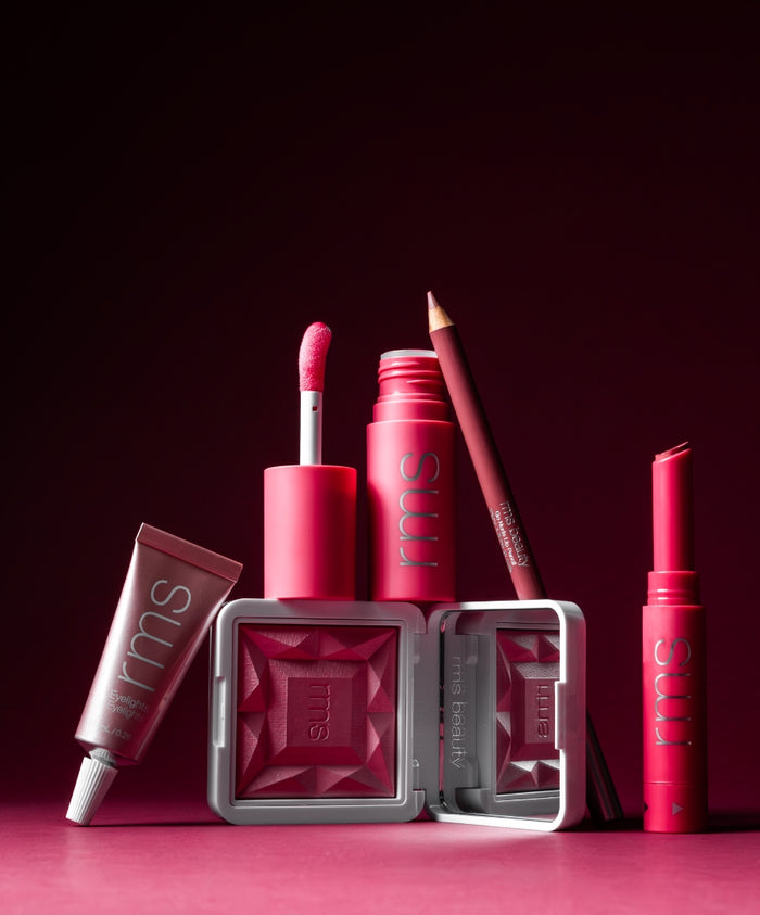 A vibrant assortment of RMS Beauty makeup products in various shades of pink and red arranged against a deep rose background, showcasing Color Capsule: The Rosé Edit with clean, cruelty-free ingredients and sustainable packaging.