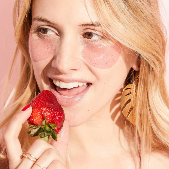 Woman wearing pink eye gels while holding a fresh strawberry against a peach background.