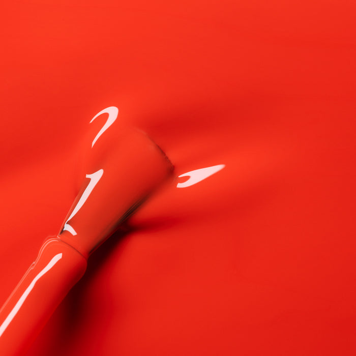 A close-up of a brush applying Manucurist's Coral Reef, a glossy orange-red gel polish, onto a smooth surface with reflections of light on the wet finish.
