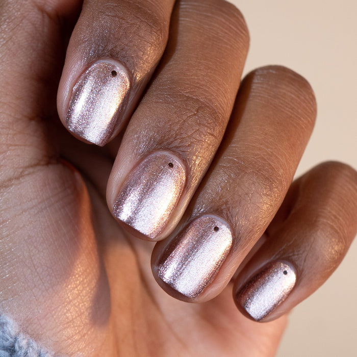 A medium-dark hand showcases Manucurist’s Cosmic Rose, an iridescent pink polish, with each nail adorned by a small black dot near the base. The background is neutral.