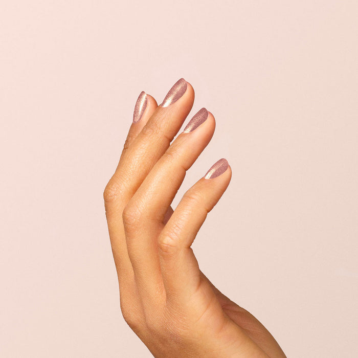 A hand with medium brown skin is shown against a pale background, fingers gracefully bent to display neatly manicured nails painted with Manucurist Cosmic Rose, an iridescent pink nail polish.