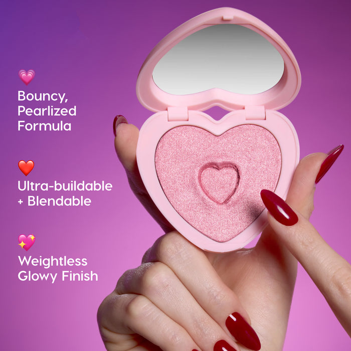 Hand with glossy red nails taps the shimmery pink Not Your Baby Super Shock Blush in a heart-shaped compact, highlighting Heart to Get bouncy pearlized formula, ultra-buildable blendability, and weightless glowy finish against a purple background.