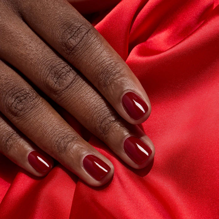 A close-up of a hand with neatly manicured nails painted in Dark Pansy by Manucurist, resting on vibrant red satin fabric.