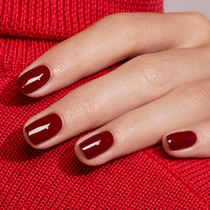 A hand with neatly manicured nails painted in Manucurist’s Dark Pansy, a glossy burgundy shade, rests on red knit fabric, perfectly matching the velvet red sweater underneath.