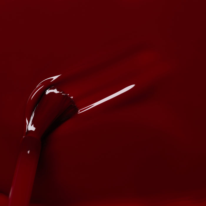 A close-up of a brush applying Manucurist's Dark Pansy nail polish, revealing glossy, smooth red strokes on a deep crimson background.