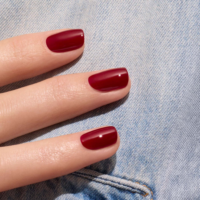 A hand with neatly manicured nails painted in Manucurist's Dark Pansy, a glossy velvet red shade, rests on light blue denim fabric.