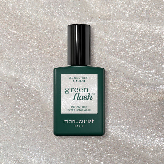 A bottle of Manucurist Diamant gel polish rests on a sparkly, light surface. The dark green bottle with a black cap and white label showcases the shimmering silver nail color.