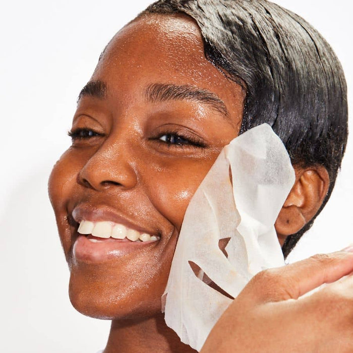 Woman removing white sheet face mask, revealing glowing skin during hydrating skincare routine