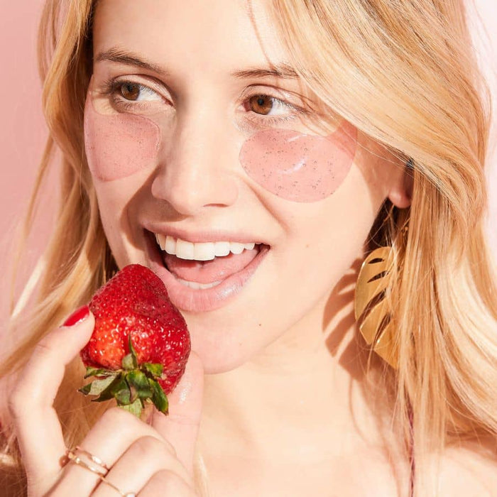Smiling blonde woman holding a fresh strawberry and wearing Patchology Rosé All Day eye gels, featuring pink, speckled hydrogel patches designed to hydrate and brighten the under-eye area.