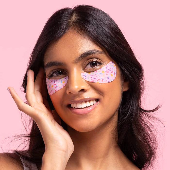 Smiling woman wearing sprinkle-pattern under-eye gel patches on both eyes, touching her face against a soft pink background