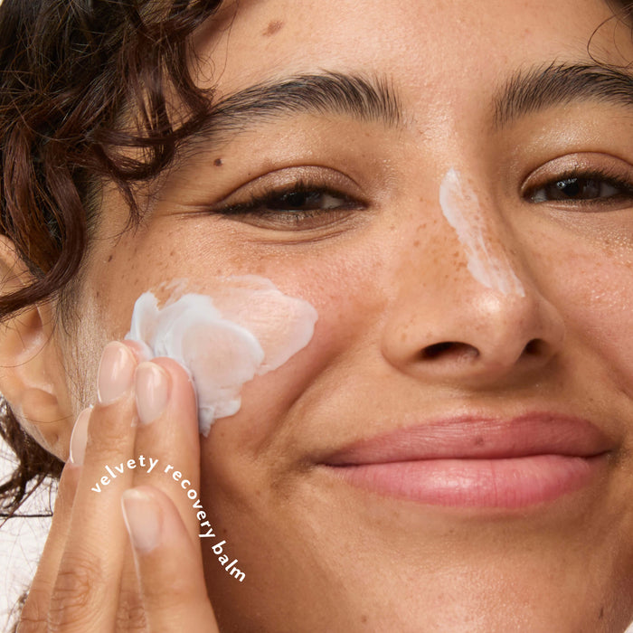 Close-up of a person applying a white five-to-nine cream balm to their cheek, with another balm patch visible on their nose. The text 'velvet recovery balm' is overlaid on the hand.