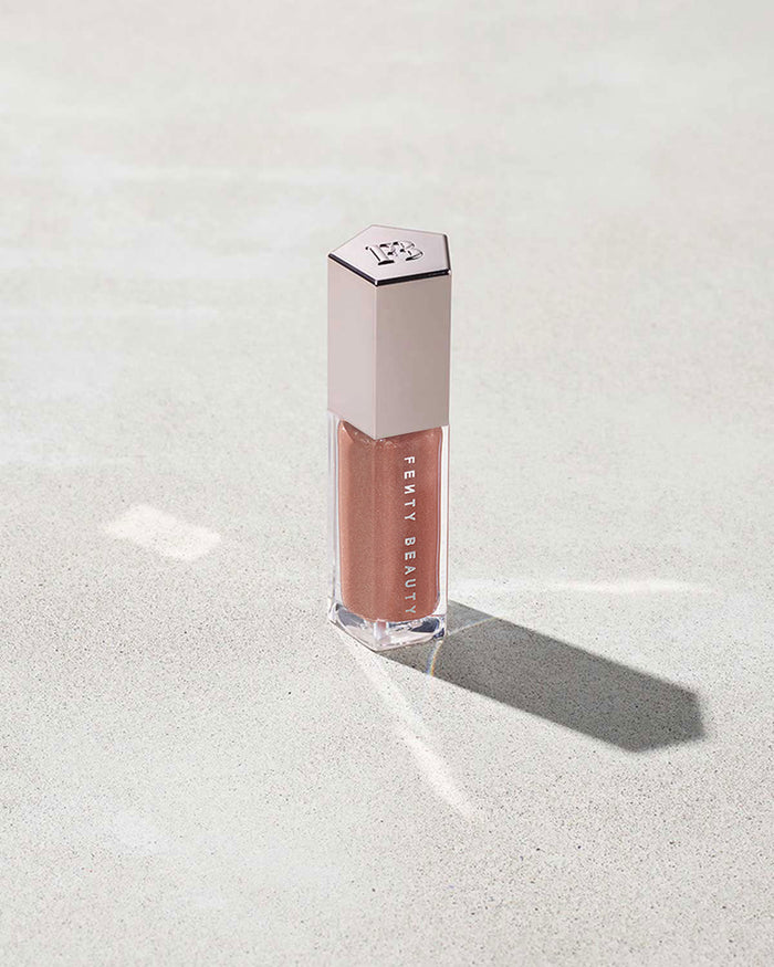 A closed tube of Gloss Bomb Universal Lip Luminizer in the shade Fenty Glow on a grey background.