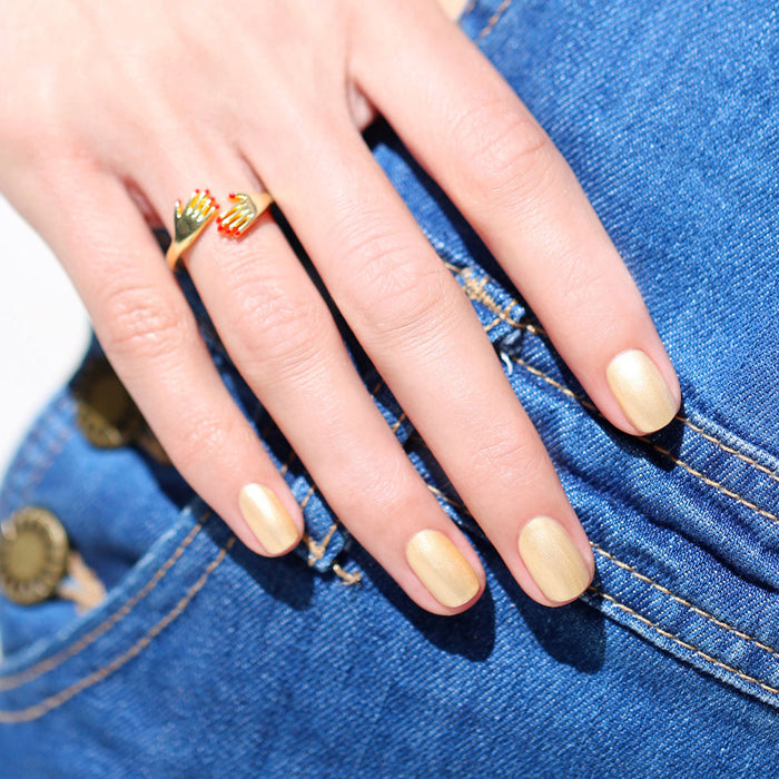 A hand with Manucurist's Gold Sand iridescent nail polish and a gold ring rests on blue denim jeans near a pocket.