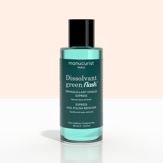 A green bottle of + Green Flash™ Gel Polish Remover by Manucurist-test, gentle and acetone-free with 99.4% bio-sourced ingredients, stands on a pale surface against a light background.