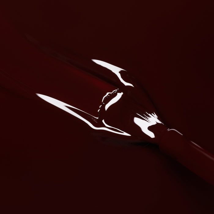 A close-up of Manucurist's Hollyhock gel polish shows its glossy red finish, smooth reflective surface, and visible brush stroke against a deep red background.