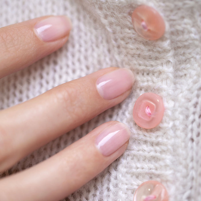 A close-up of a hand with neatly manicured nails painted in Manucurist's Hortencia, a sheer pink gel polish, gently touching a white knitted fabric with pink buttons.