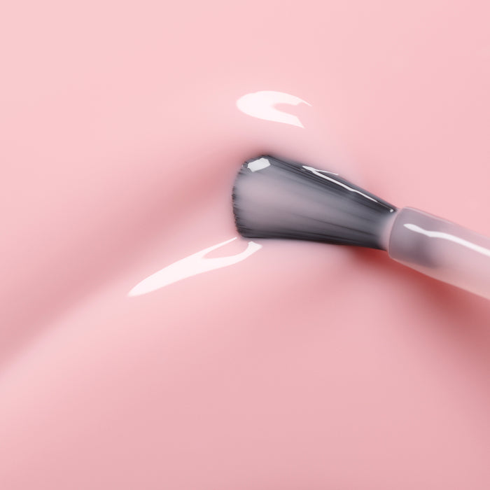 A close-up of a Manucurist Hortencia nail polish brush applying sheer pink gel polish onto a smooth pink surface with shiny highlights.