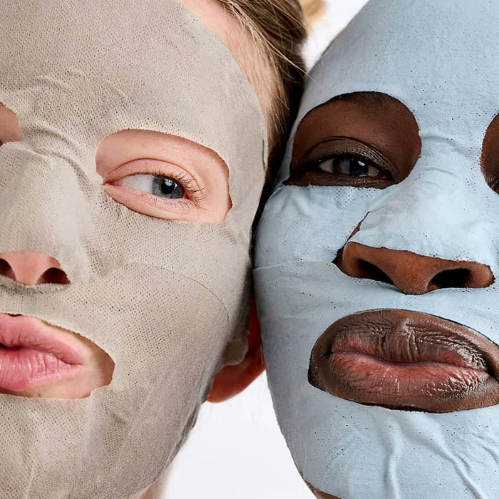 Two women wearing grey and blue clay sheet masks, close-up skincare treatment faces touching together
