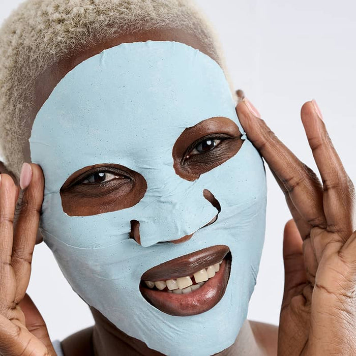 Woman wearing Patchology Mudmask Hydrate smiling and holding mask edges, hydrating skincare treatment close-up