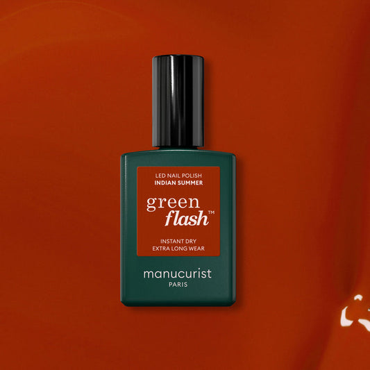 A bottle of Manucurist Indian Summer Green Flash LED nail polish—an ochre red shade—stands against a matching orange-red background, with a dark green bottle, black cap, and white text.