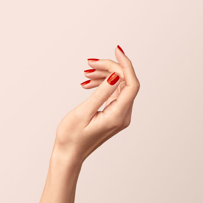 A fair-skinned hand with neatly manicured nails painted in glossy "Indian Summer" by Manucurist is shown against a light beige background. The fingers are gently curved in a relaxed pose, highlighting the vibrant brick red gel polish.