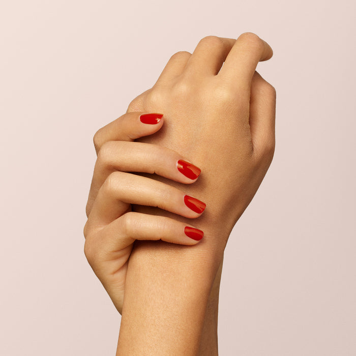Two hands are shown against a plain, light background. One gently holds the wrist of the other, showcasing neatly manicured nails painted with Manucurist's Indian Summer, a glossy brick red gel polish.