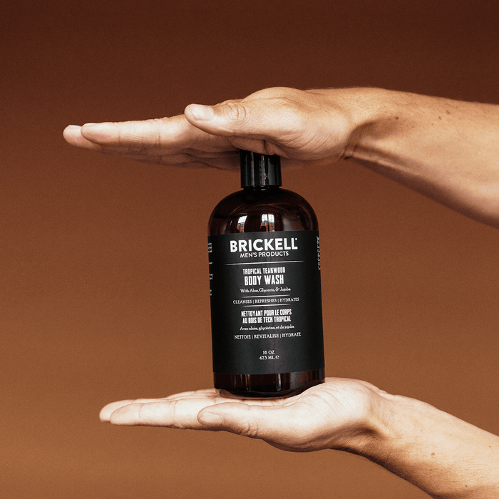 Brickell Men's Products Tropical Teakwood Body Wash bottle held between two hands with brown background