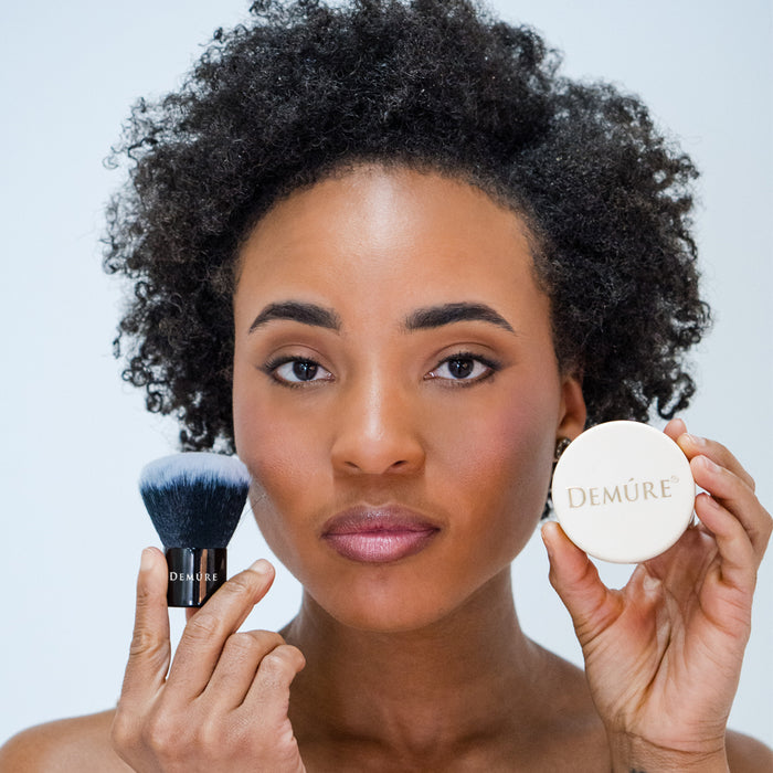 Woman's face with her holding a Demure Kabuki brush in one hand and a jar of Demure Foundation makeup in the other hand.