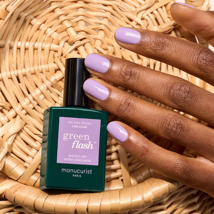 A hand with neatly manicured, light purple nails rests on a woven basket beside a Manucurist Lisa Lilas bottle, adding chic 90s vibes.