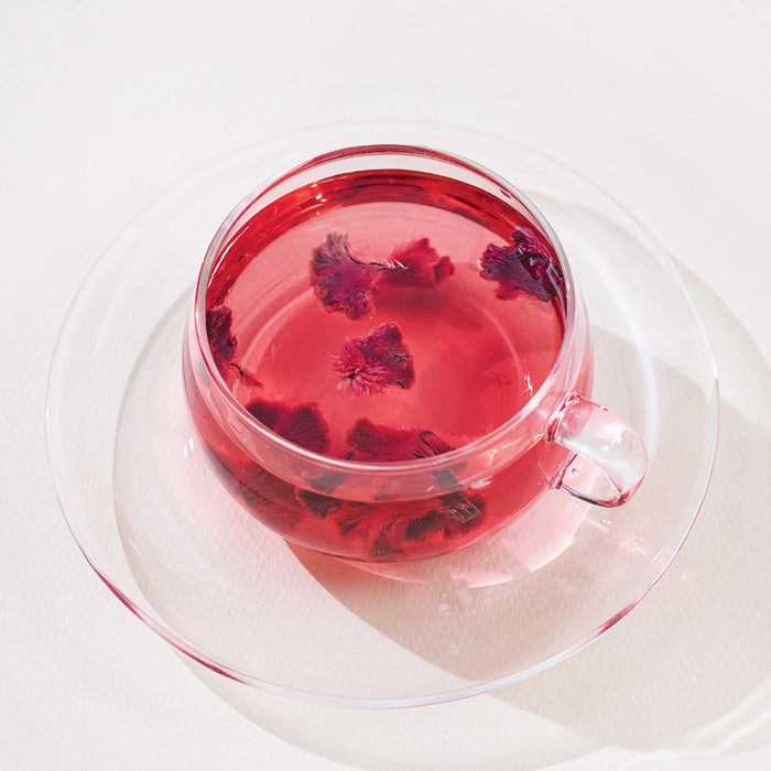 Clear glass mug of pink flower tea with visible dried blossoms, isolated view